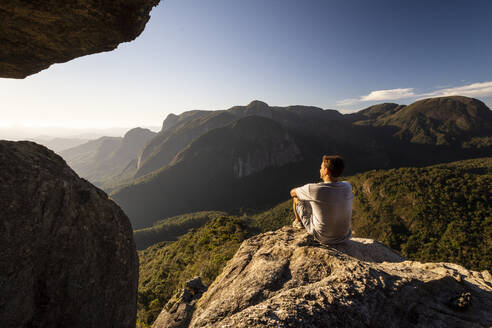 Man sitting on rocky edge looking at beautiful view to mountains - CAVF73024