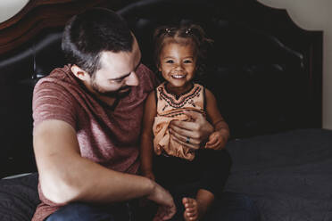 Beautiful smiling 2 yr old multiracial girl sitting on bed with dad - CAVF73032