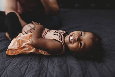 Süßes multirassisches 2 Jahre altes Mädchen liegt lachend auf dem Bett - CAVF73033