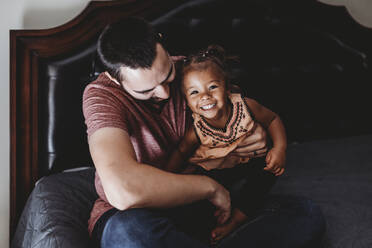 Dark haired dad sitting on bed with smiling multiracial 2 yr old - CAVF73035