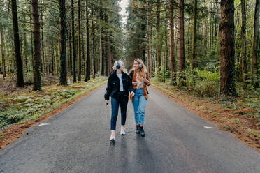 Two women walking on a road in the forest while listening to music - CAVF73089