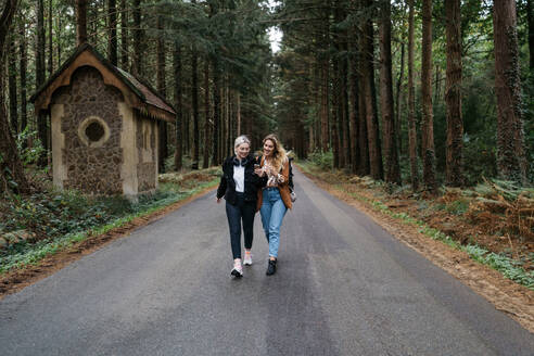 Zwei Frauen, die auf einer Straße im Wald spazieren gehen und dabei auf ihr Handy schauen - CAVF73090