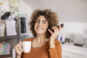 Frau hält Mobiltelefon und Kaffeetasse zu Hause - FMKF06073