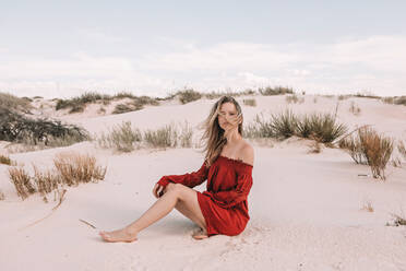 Eine Frau in den Zwanzigern sitzt in einem roten Kleid auf dem Sand - CAVF73276