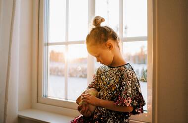 Junges Mädchen kuschelt mit ihrem Spielzeug in einem glitzernden Kleid zu Hause bei Sonnenuntergang - CAVF73337