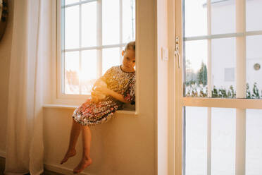 Junges Mädchen, das traurig auf einer Fensterbank zu Hause bei Sonnenuntergang sitzt - CAVF73340