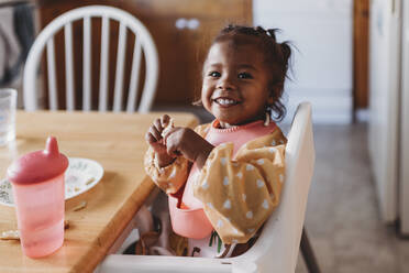 Lächelndes 2-jähriges gemischtrassiges Mädchen mit Lätzchen im Hochstuhl in der Küche - CAVF73429