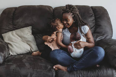 Multirassische 2 Jahre alte küssende Mutter mit Zöpfen, die ihr Neugeborenes auf der Couch hält - CAVF73434