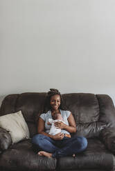 Lächelnde ethnische Mutter hält Neugeborenes auf Ledercouch unter Wandkunst - CAVF73437