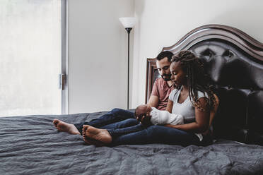 Multiracial couple sitting on bed admiring their newborn baby - CAVF73450