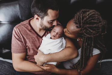 Multiracial parents kissing newborn baby wrapped in white blanket - CAVF73452