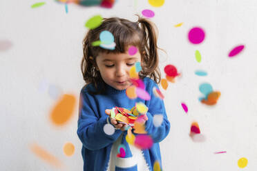 Happy little girl blowing the confetti at a party in front of a white wall - GEMF03412