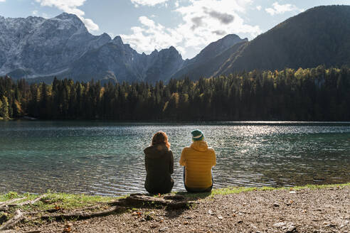 Rear view of couple sitting on lakeside at Laghi di Fusine, Friuli Venezia Giulia, Italy - MAUF03225