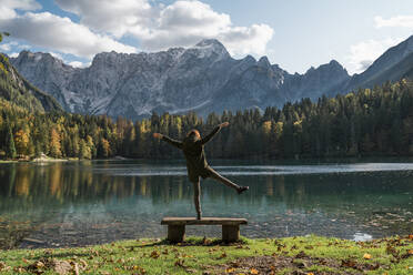 Rear view of female hiker standing on one leg on a bench at Laghi di Fusine, Friuli Venezia Giulia, Italy - MAUF03233