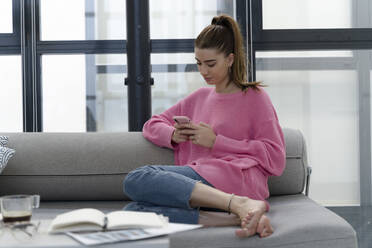 Junge Frau sitzt barfuß auf der Couch und benutzt ein Mobiltelefon - ERRF02570