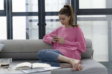 Young woman sitting barefoot on the couch using cell phone - ERRF02570