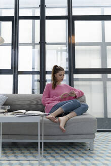 Junge Frau sitzt barfuß auf der Couch und benutzt ein Mobiltelefon - ERRF02571
