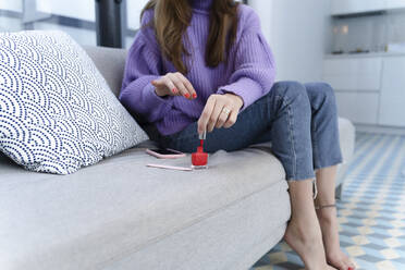 Crop view of young woman sitting on couch applying nail varnish - ERRF02590