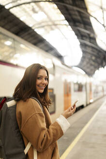 Porträt einer lächelnden jungen Frau mit Mobiltelefon am Bahnhof - VABF02493