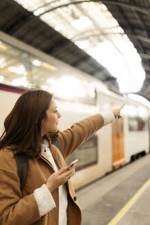 Junge Frau mit Handy auf dem Bahnhof zeigt mit dem Finger - VABF02494