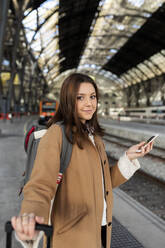 Porträt einer lächelnden jungen Frau mit einem Handy in der Hand am Bahnhof - VABF02501