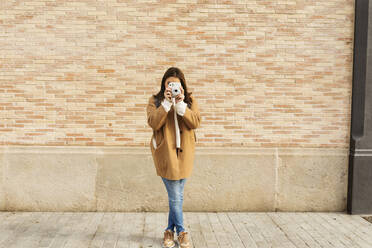 Junge Frau mit Kamera, die vor einer Backsteinmauer fotografiert - VABF02520