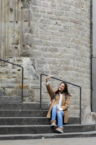 Lächelnde junge Frau sitzt auf einer Treppe in der Stadt und macht ein Selfie, Barcelona, Spanien, lizenzfreies Stockfoto