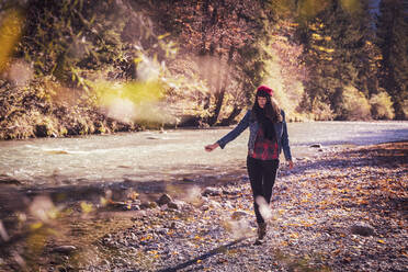 Woman wearing red woolly hat and denim jacket walking at riverside in autumn - DHEF00020
