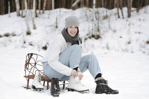 Porträt einer lächelnden Frau, die Schlittschuhe auf einem Schneefeld anzieht - EYAF00813