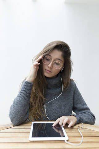 Porträt einer jungen Frau, die an einem Holztisch sitzt und Kopfhörer und ein digitales Tablet benutzt, lizenzfreies Stockfoto