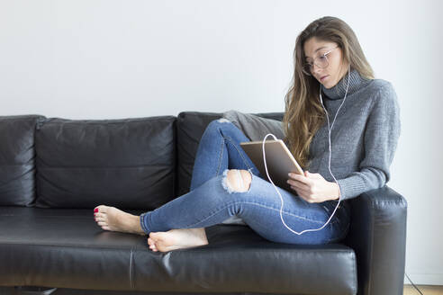 Junge Frau sitzt auf einer schwarzen Ledercouch mit Kopfhörern und digitalem Tablet - JPTF00425