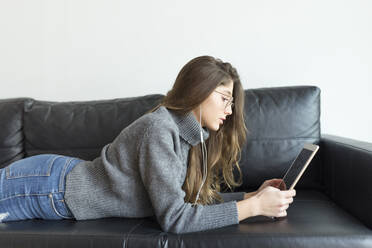 Young woman lying on black leather couch using earphones and digital tablet - JPTF00426