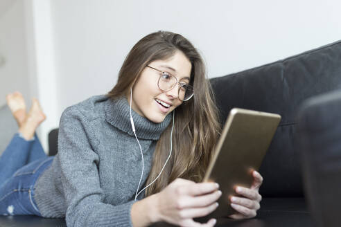 Porträt einer lächelnden jungen Frau, die mit Kopfhörern und digitalem Tablet auf der Couch liegt - JPTF00427