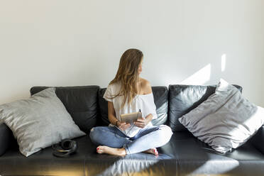 Junge Frau sitzt mit Kopfhörern und digitalem Tablet auf der Couch - JPTF00433