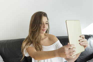 Porträt einer jungen Frau, die auf einer Couch sitzt und ein Selfie mit einem digitalen Tablet macht - JPTF00434