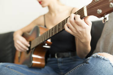 Crop view of young woman sitting on couch playing guitar - JPTF00448