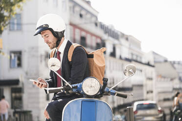 Young businessman on motor scooter using cell phone in the city, Lisbon, Portugal - UUF20072