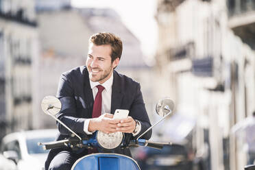 Young businessman with cell phone on motor scooter in the city, Lisbon, Portugal - UUF20085