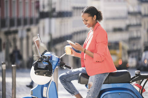 Lächelnde junge Frau, die ein Mobiltelefon auf einem Motorroller in der Stadt benutzt, Lissabon, Portugal, lizenzfreies Stockfoto