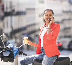 Smiling young woman with motor scooter talking on the phone in the city, Lisbon, Portugal - UUF20092