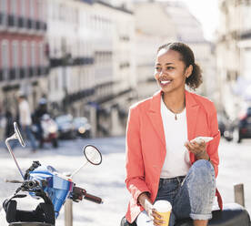 Smiling young woman with motor scooter and cell phone in the city, Lisbon, Portugal - UUF20097