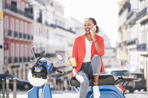 Laughing young woman with motor scooter talking on the phone in the city, Lisbon, Portugal - UUF20098