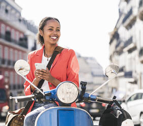 Lächelnde junge Frau mit Motorroller und Mobiltelefon in der Stadt, Lissabon, Portugal - UUF20107