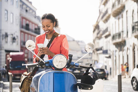 Lächelnde junge Frau, die ein Mobiltelefon auf einem Motorroller in der Stadt benutzt, Lissabon, Portugal, lizenzfreies Stockfoto