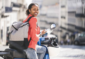 Porträt einer glücklichen jungen Frau mit Motorroller und Mobiltelefon in der Stadt, Lissabon, Portugal - UUF20109