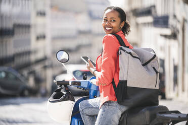 Smiling young woman with motor scooter and cell phone in the city, Lisbon, Portugal - UUF20110