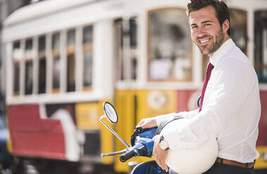 Portrait of smiling young businessman on motor scooter in the city, Lisbon, Portugal - UUF20113