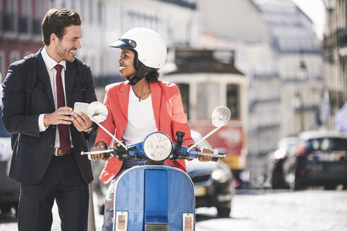 Glückliches junges Geschäftspaar mit Mobiltelefon und Motorroller in der Stadt, Lissabon, Portugal - UUF20122