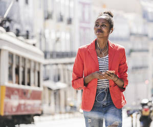 Young woman with earphones and smartphone looking around in the city, Lisbon, Portugal - UUF20133