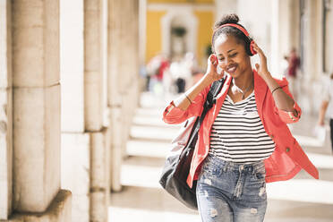 Glückliche junge Frau mit Kopfhörern beim Musikhören in der Stadt, Lissabon, Portugal - UUF20136